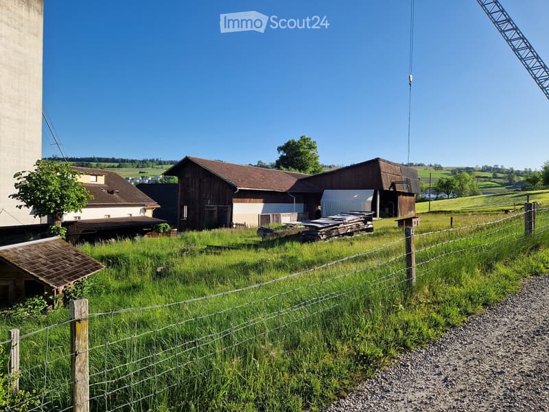A farm house with brown roof and a barn, surrounded by a fenced yard and green grass.