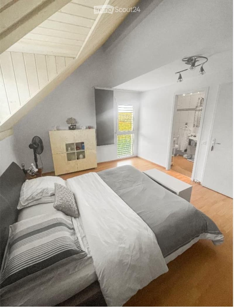 A bedroom with a bed, white bedding, and pillows, wooden floor, beige cabinet with glass doors, fan, white painted walls, wooden ceiling, large window with white frame and shutters, mirror, open door leading to a bathroom.
