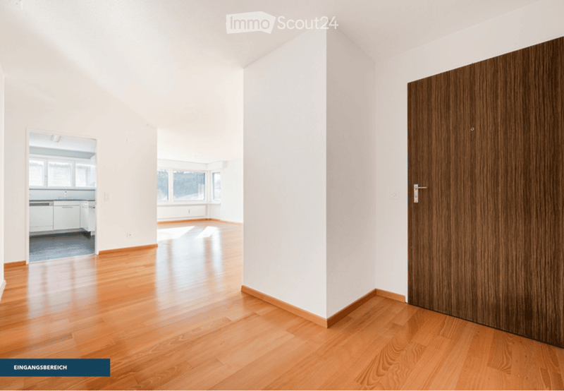 Empty entrance area with light wooden flooring, white walls, and a closed wooden door. The space is clean and unfurnished, with a view into an adjacent kitchen area through an open doorway.