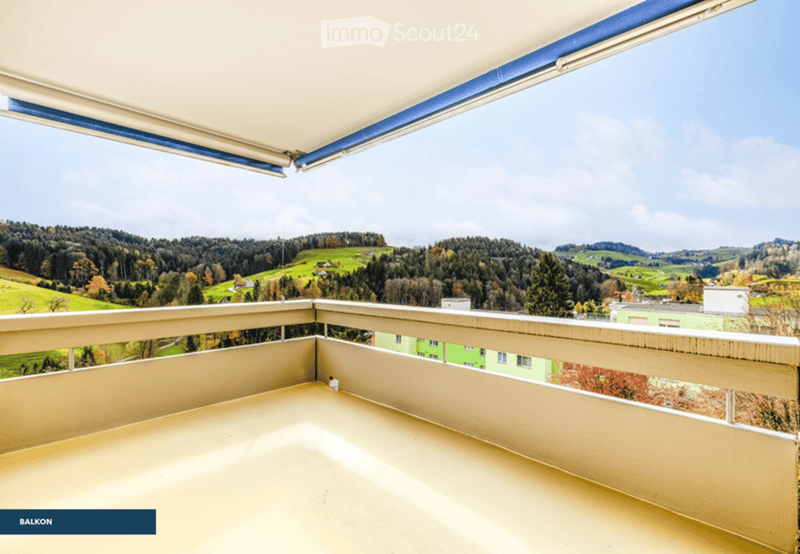 Empty balcony with wooden railing and a retractable awning, offering a scenic view of a green valley and forested hills.