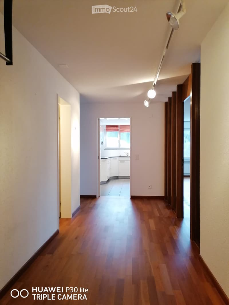 Empty hallway with wooden floor, white walls, track lighting, open door to kitchen