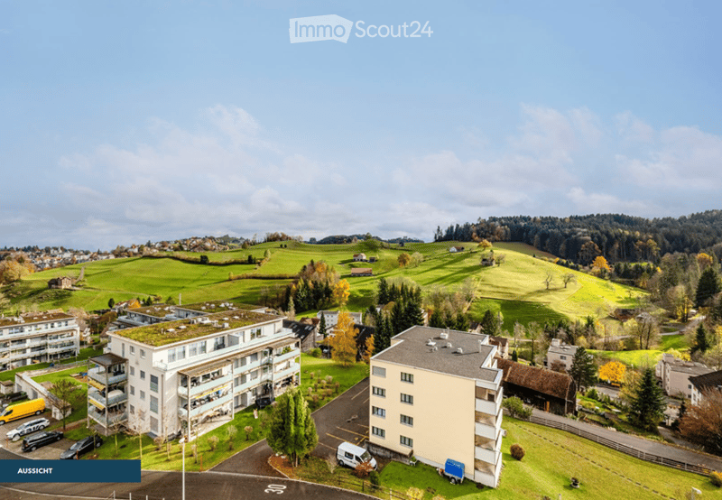 Aerial view of residential buildings, green fields, trees, and parked vehicles in a scenic landscape.