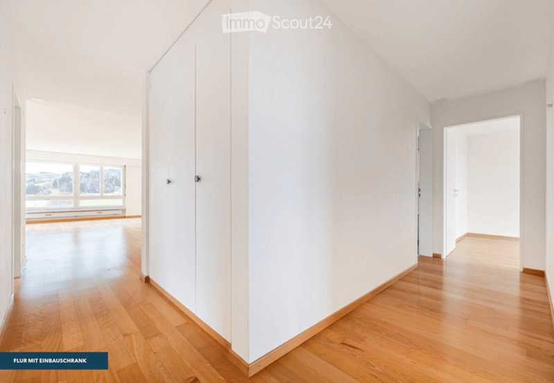 Empty hallway with light wooden flooring and white walls, featuring a large window at one end and an open doorway leading to another room.