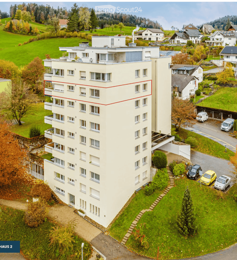 Aerial view of a multi-story residential building with balconies, surrounded by greenery and parked cars.