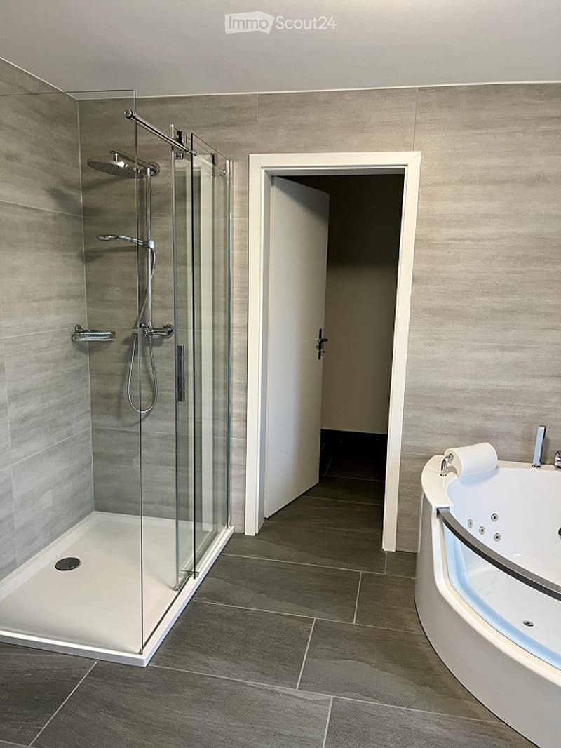 A modern bathroom with a glass shower cubicle, large white bathtub, tiled flooring, and tiled walls.