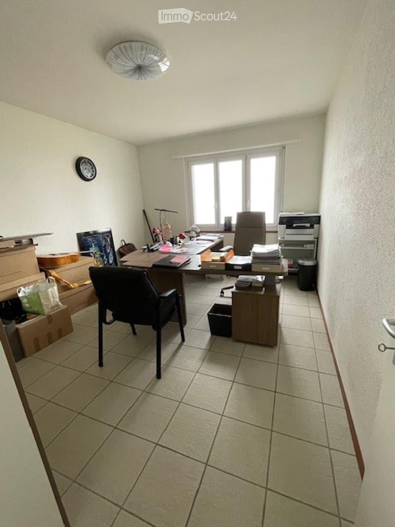 The office has a desk, two chairs, a printer, and a trash can. The room also contains some boxes, bags, and miscellaneous items.