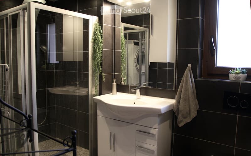 modern bathroom with white sink and shower, black tiles, towel rack with towel, plant on the window sill, washing machine behind mirror