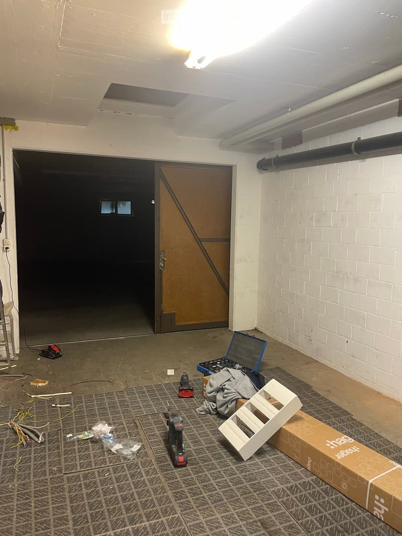 Empty room with exposed brick wall, concrete floor, and two doors, one leading to a dark space. Tools and construction materials are scattered on the floor.