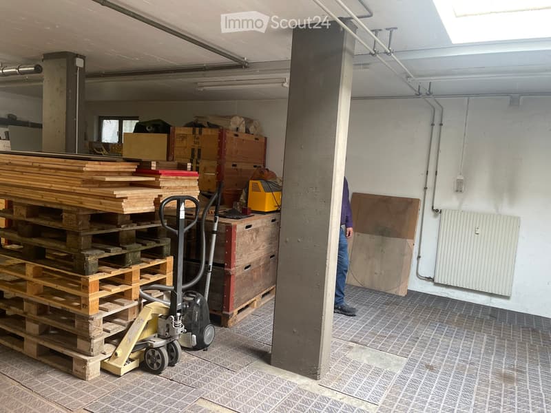 A spacious storage area with stacked pallets, wooden planks, and a vacuum cleaner. The space features exposed concrete pillars, a radiator, and a tiled floor.