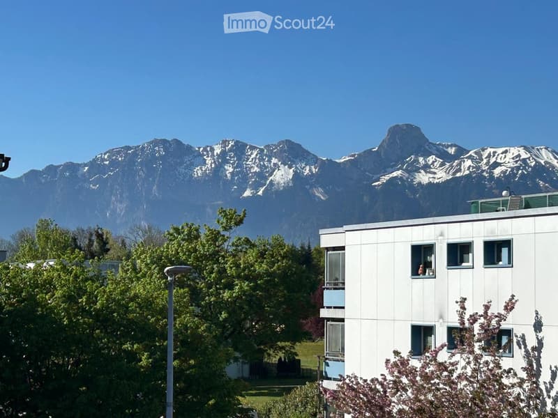 White building with multiple windows, surrounded by trees, mountains in the background