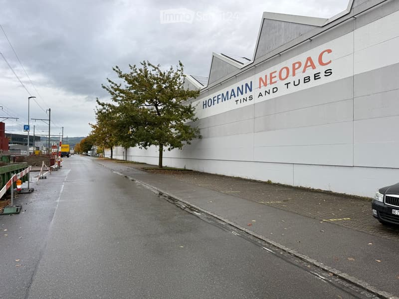An industrial building exterior, featuring a large white wall with a name and logo reading 'HOFMANN NEOPAC TINS AND TUBES'.