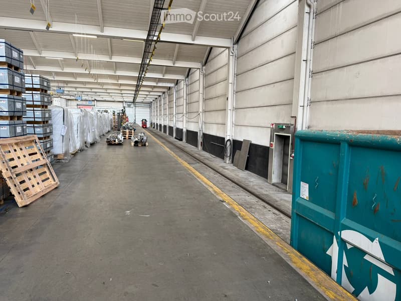 Warehouse with grey concrete flooring, high ceilings with exposed steel beams, white and grey walls, yellow demarcation line on the floor, multiple pallets and crates along the side.