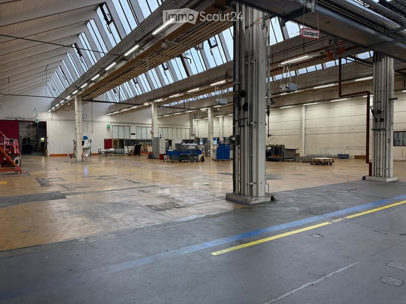 A large open space with concrete floors and a glass roof. The interior has several pillars and equipment pieces spread throughout. There are various metal structures and machinery visible, and the space is illuminated by fluorescent lights on the ceiling.