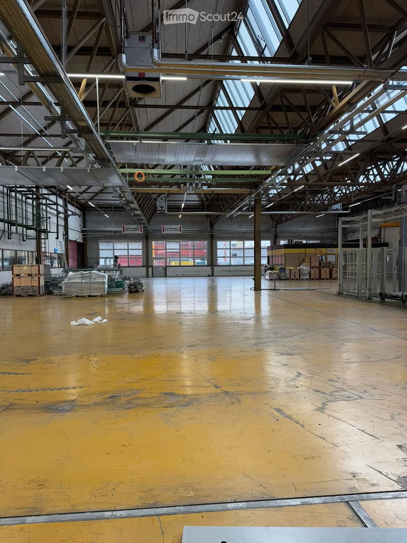 Wide warehouse space, yellow floor, metal beams, overhead lights, storage boxes, glass windows.