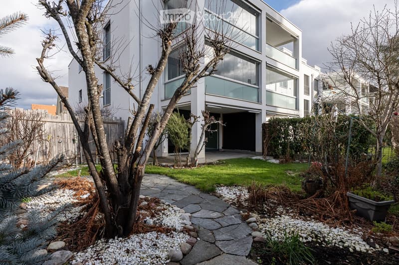 Modern two-story building with large glass windows, concrete pathway, fenced garden, shrubs, trees, driveway, garage entrance