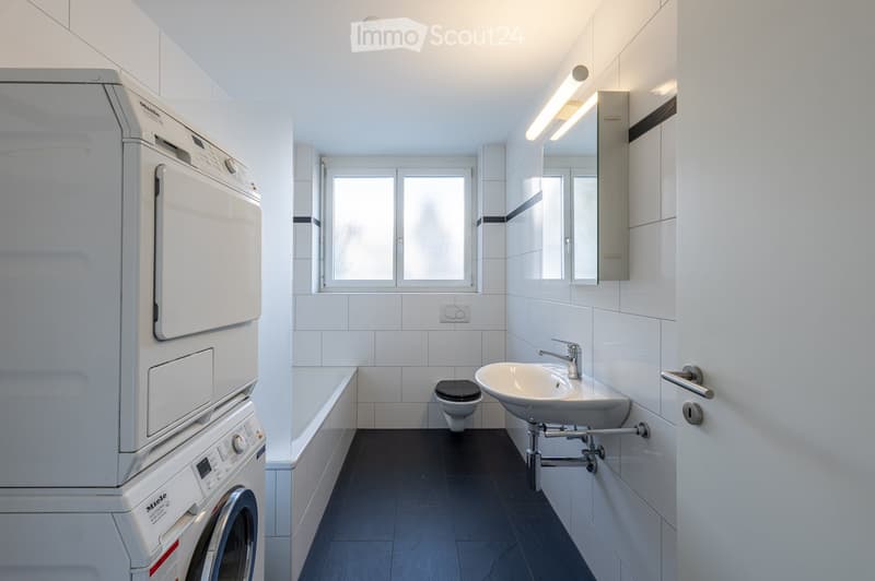 white tiled walls, white tile floor, black tile flooring, a sink, a toilet, a bathtub, a stacked washer and dryer unit