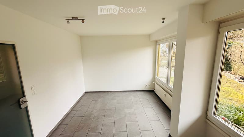 Empty room with white walls, tiled floor, window with yellow trim, ceiling lights, power outlets, and a door.
