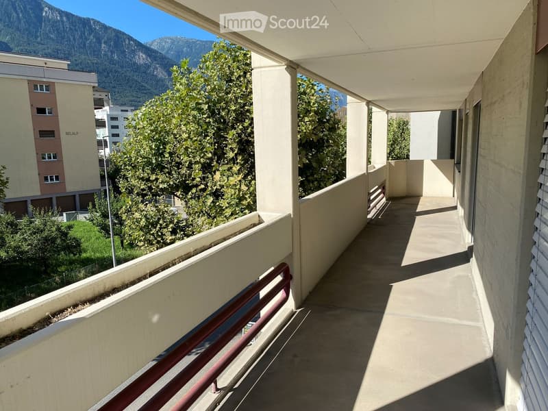 balcony with red railing, concrete floor, shadow of balcony on the floor, greenery, mountains in the background