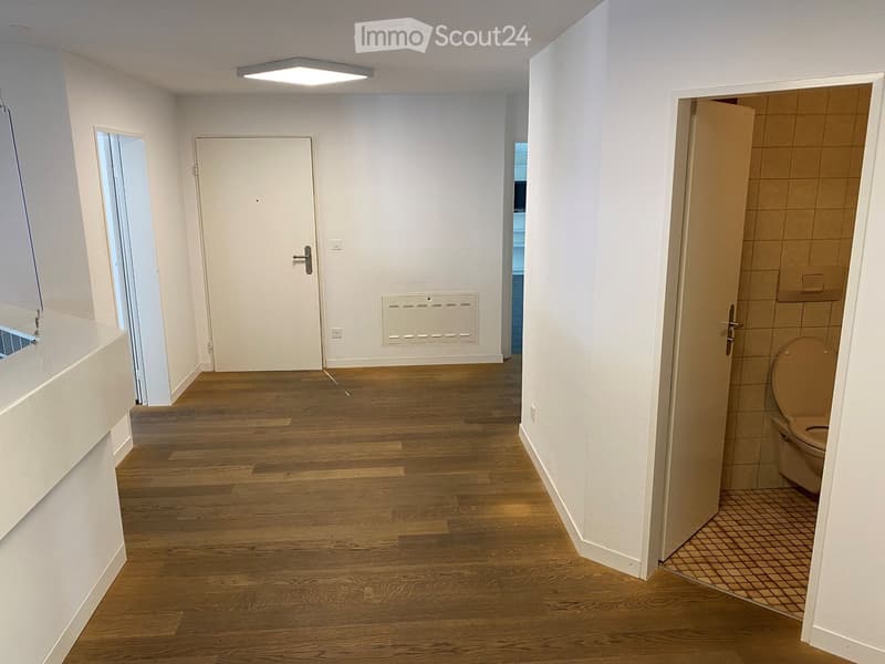 Empty room with hardwood floors, white walls, ceiling light, open door to another room, white door, radiator, closed door to the bathroom with toilet visible