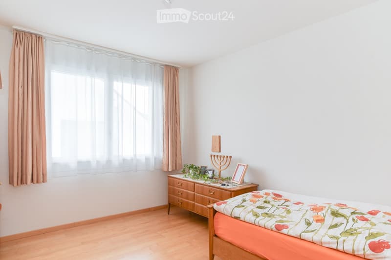 Small bedroom with wooden floor, white walls, large window, wooden cabinet, and floral bedspread.