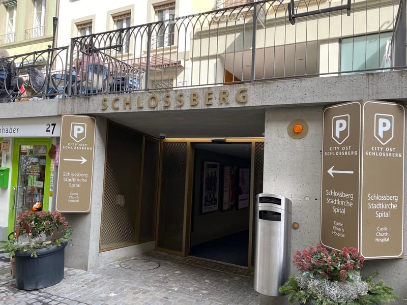 Building exterior of Schlossberg City Ost, entrance, two signboards, trash can, two potted plants, brick floor