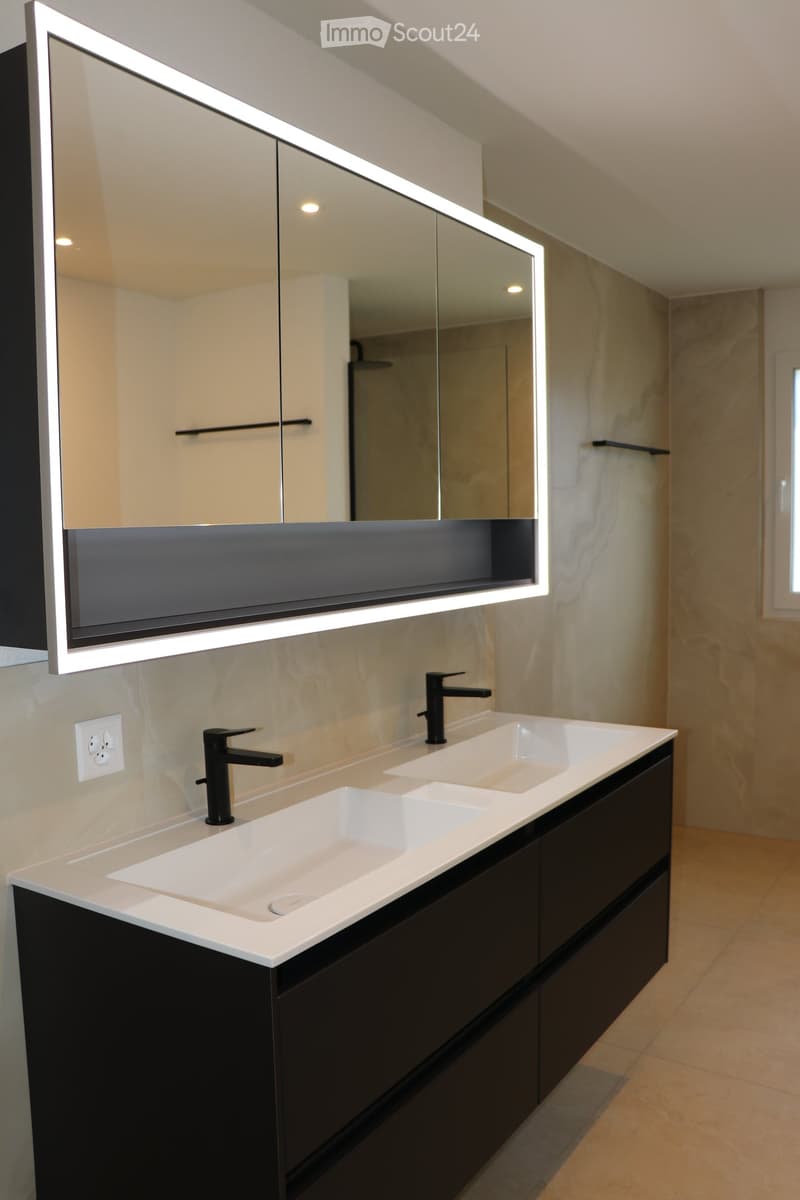 modern bathroom with double sink vanity, black cabinet, large mirror with integrated lighting, white tiled floor, silver faucets, power outlet