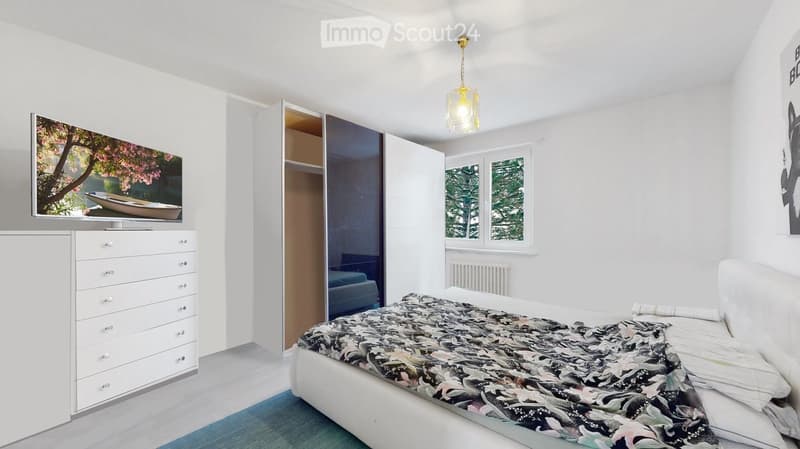 A white bedroom with a white bed, a blue rug, a white wardrobe, a drawer chest, a mounted television on the wall, a ceiling lamp, a window with a radiator below it, and an indoor view of a tree.