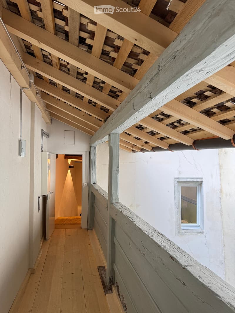 Corridor / Hallway. Wooden staircase with exposed beams and a small window, leading to a warmly lit room with wooden flooring.