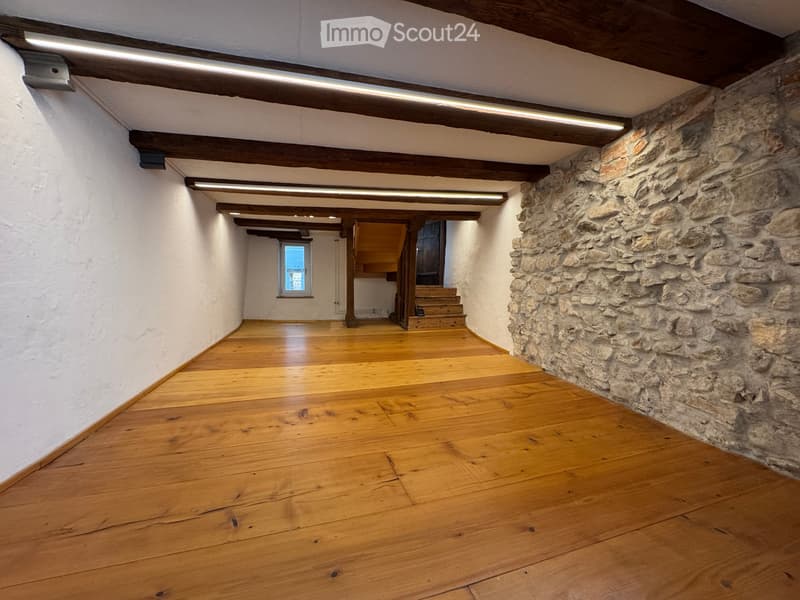 Unfurnished Room. Empty room with wooden flooring, white walls, exposed wooden beams, and a stone wall on one side. A small window and a staircase lead to another area.
