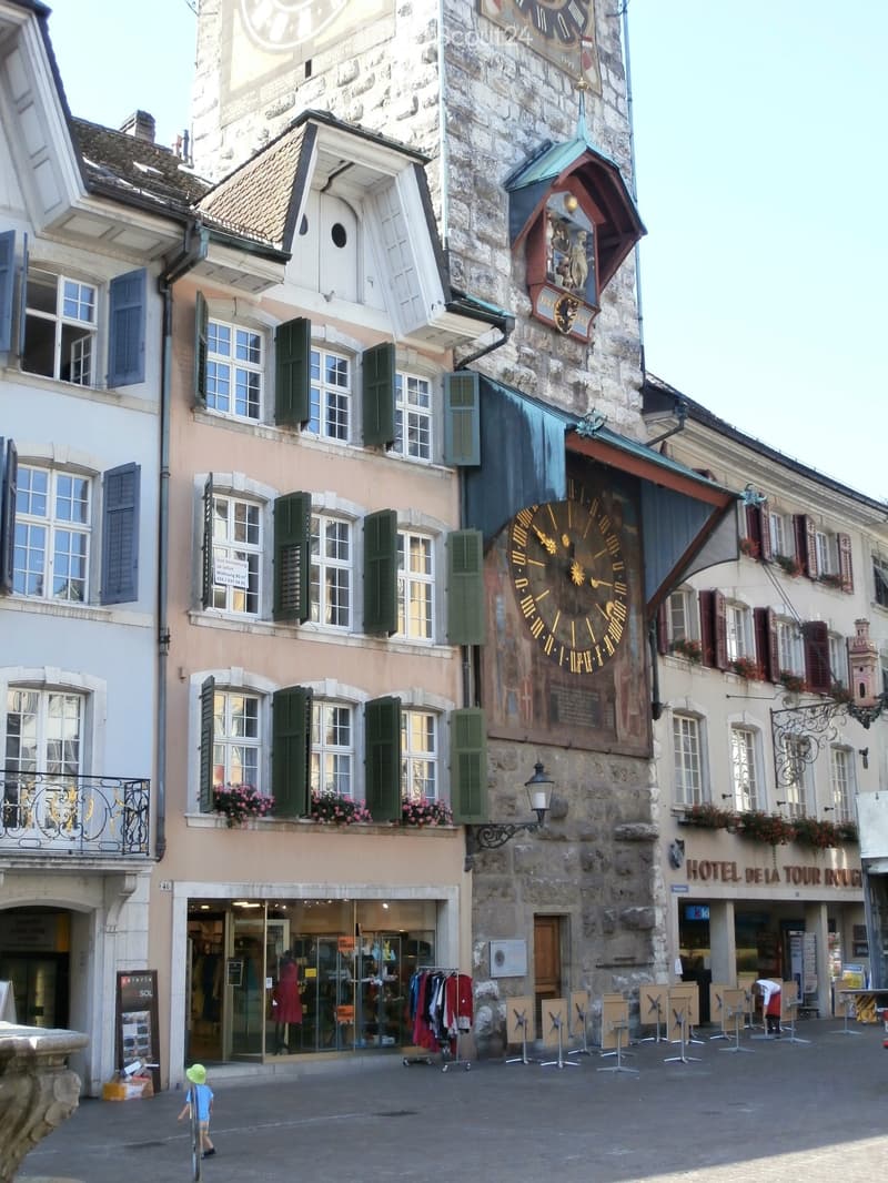 The building is three stories with stone walls and wooden eaves. A clock tower with a gold clock is protruding from the center of the building. There are two rows of windows on the second and third floors, each adorned with wooden shutters and a decorativ