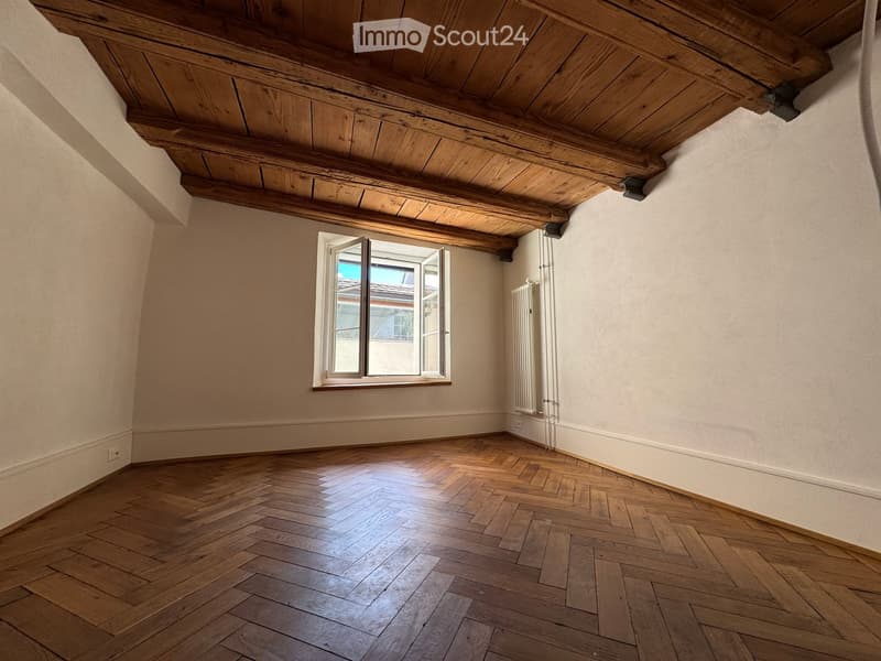 Unfurnished Room. Empty room with wooden herringbone flooring, exposed wooden ceiling beams, and a single window with white frame and radiator below.