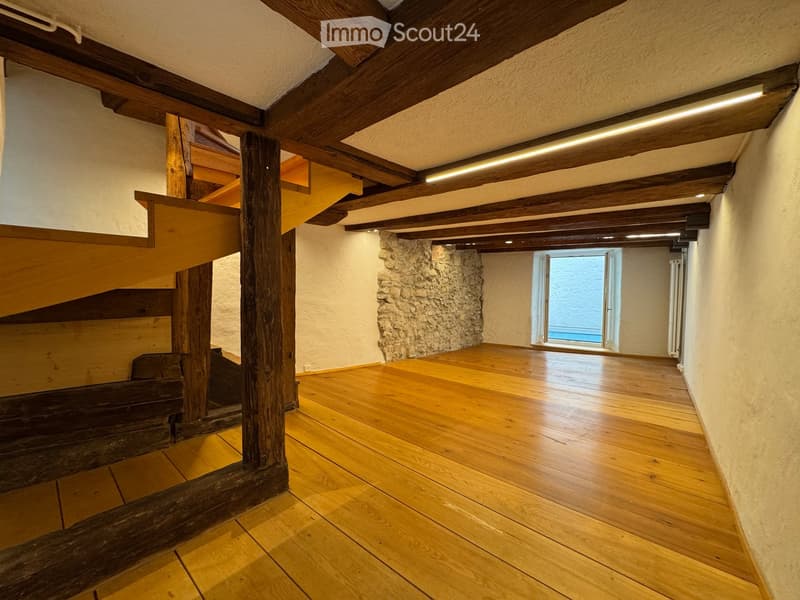 Unfurnished Room. Empty room with wooden flooring, exposed wooden beams, and a stone wall. A staircase leads upwards, and a window provides natural light.
