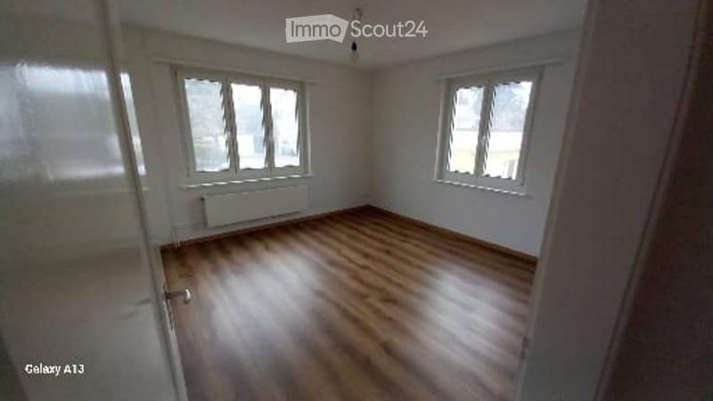 Empty room with white walls, wooden floor, radiator, two windows, light hanging from ceiling.