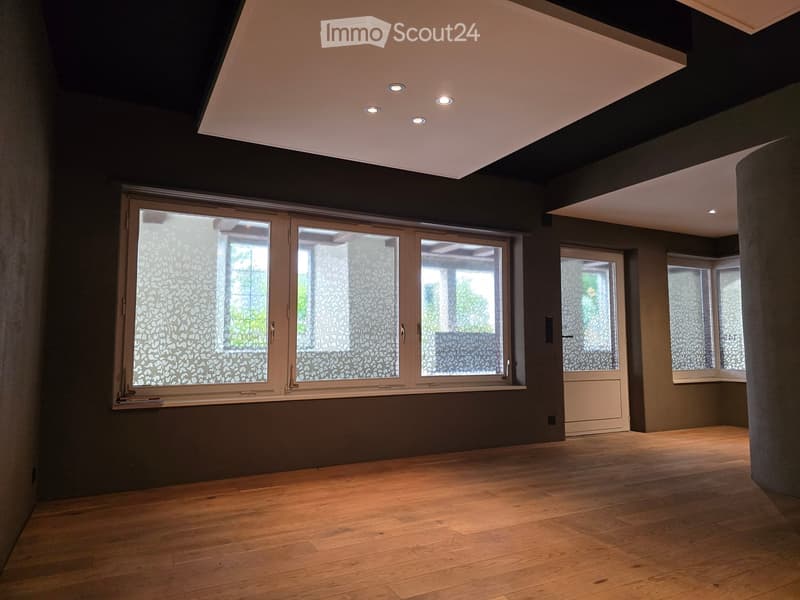 Empty room, wooden floor, grey walls, three large windows, ceiling with four small lights.