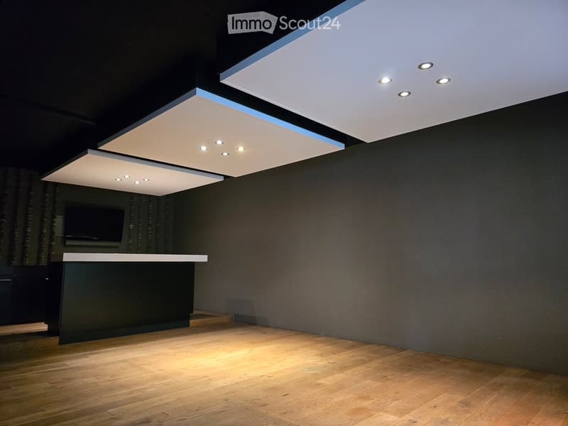 large room with exposed ceiling lights, wooden floor, black walls, counter, television mounted on wall