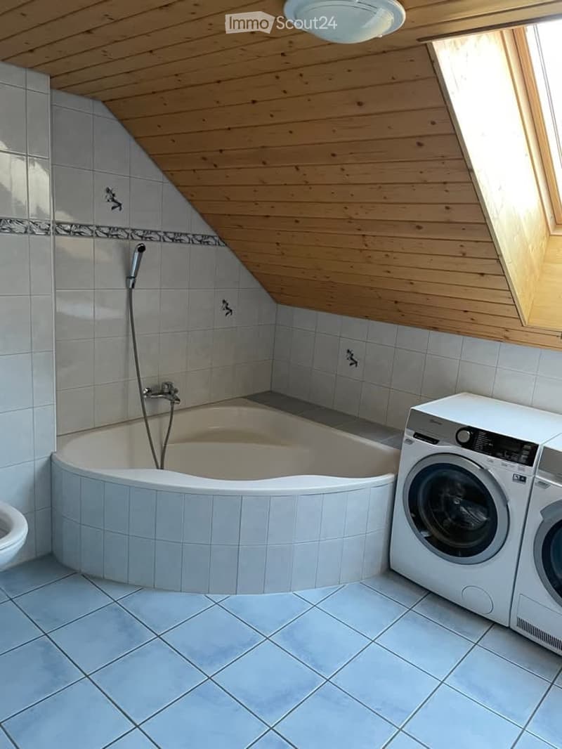 A corner bathroom with a bathtub, toilet, washing machine, tiled flooring, wooden ceiling, skylight window