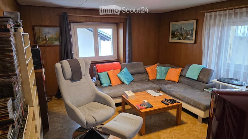 A room with wooden floor, wooden sofa with cushions, wooden table, wooden chair, desk, window with curtains, bookshelf, picture frames on the wall