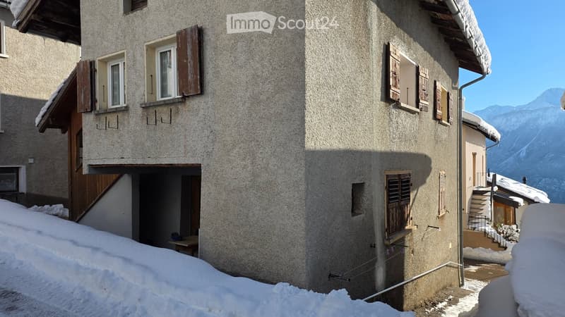 Mountainside building, stone facade, multiple windows with shutters, stairs on the right side, snow-covered ground