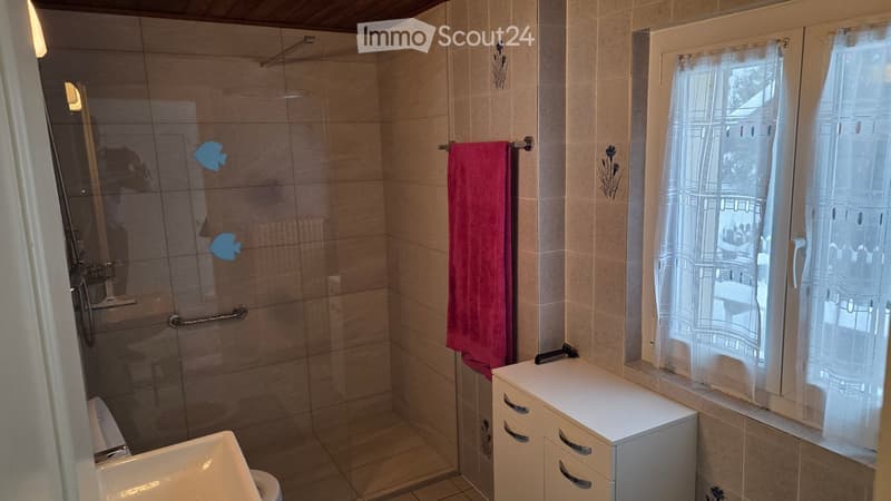 A bathroom with a toilet, a shower with glass door, a washbasin, a cabinet with drawers, a towel on the wall, and a window with curtains.