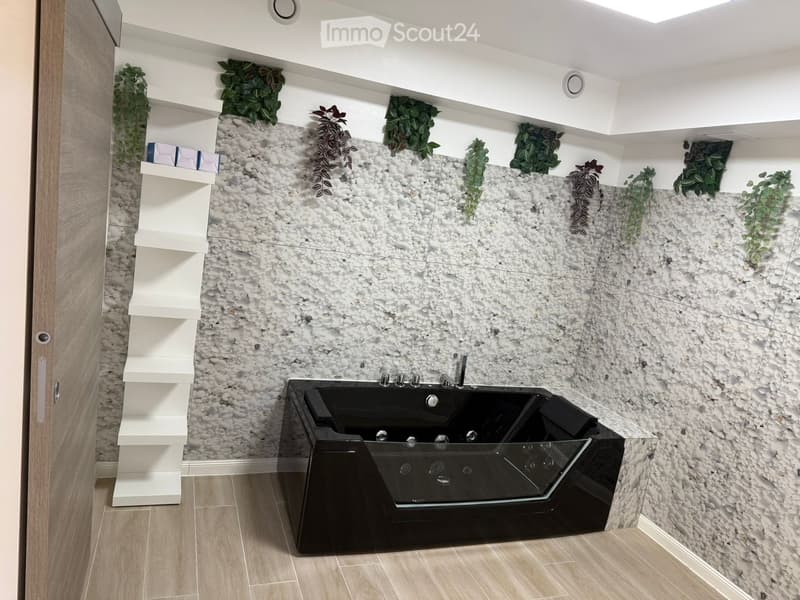 Large black tub, glass door, wooden floors, pebble-textured wall with green plants, white shelf