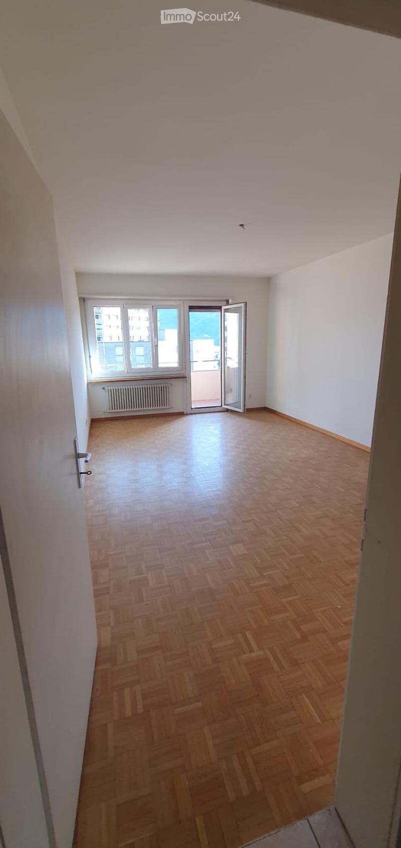 Empty room with wooden floors, sliding door leading to balcony, white walls, and large windows with a view.