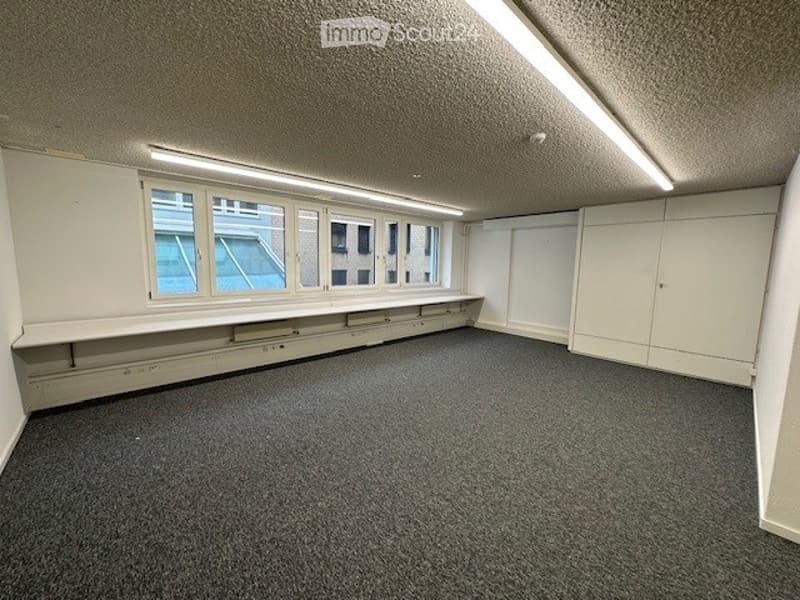 The room has white walls, a white ceiling, a white shelf, a carpeted floor, three large windows, a long counter, and a single light on the ceiling.