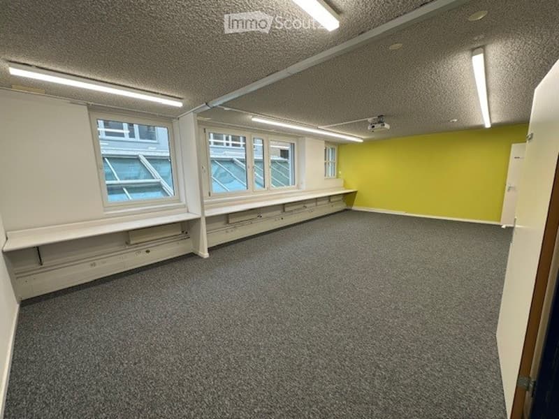 An empty room with gray carpeting, white walls, yellow walls, two windows, two benches, and fluorescent lights.