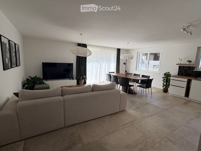 Modern living and dining area with a large sectional sofa, coffee table, dining table with chairs, television, pendant lights, and plants.