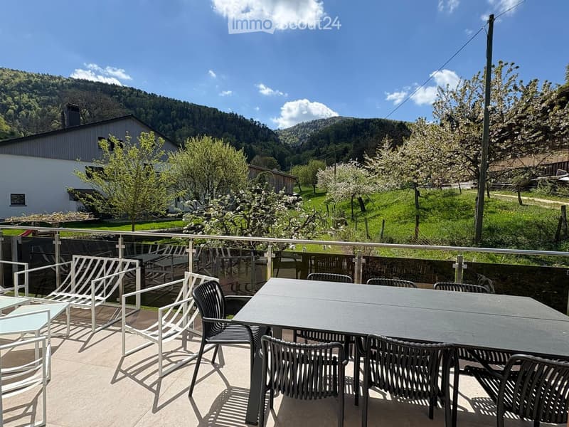 Outdoor dining area with a table and chairs, surrounded by a metal balustrade, offering a view of a house and a mountainous landscape.