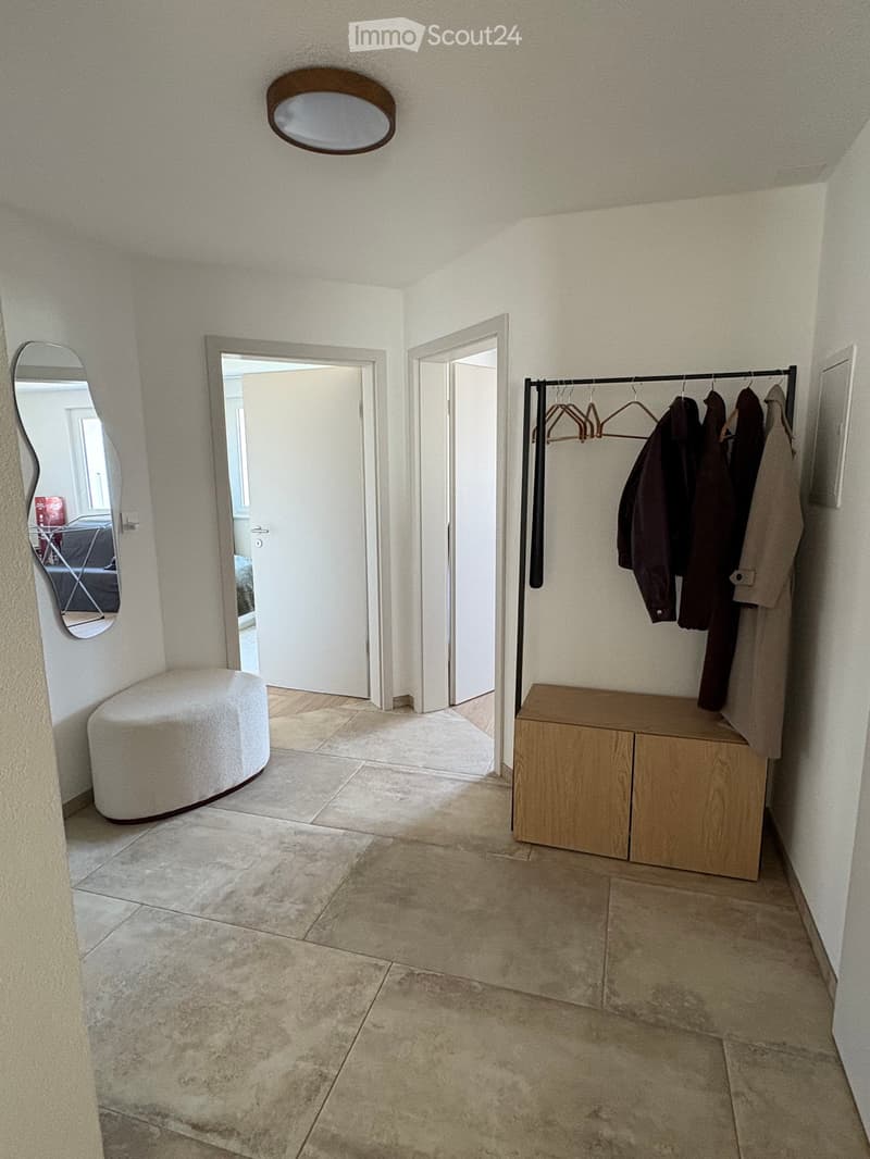 Empty hallway with tiled floor, white walls, coat rack with jackets, small bench, and two open doors leading to other rooms.