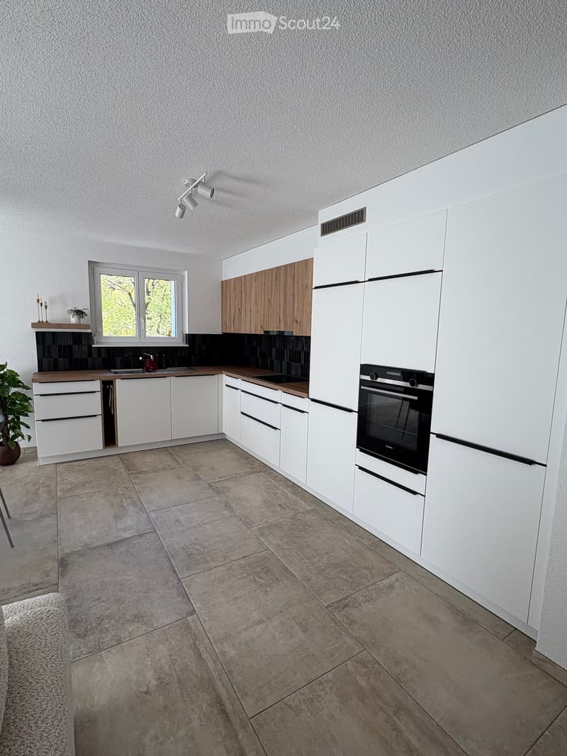 Modern kitchen with white cabinets, wooden accents, tiled floor, and built-in oven. Features a window, sink, and small plant on the counter.