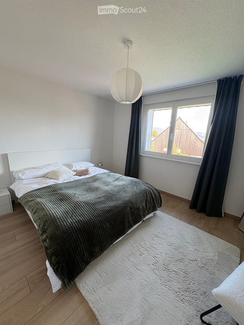 Modern bedroom with a large bed, nightstand, and large window with dark curtains. The room features wooden flooring and a hanging spherical lamp.