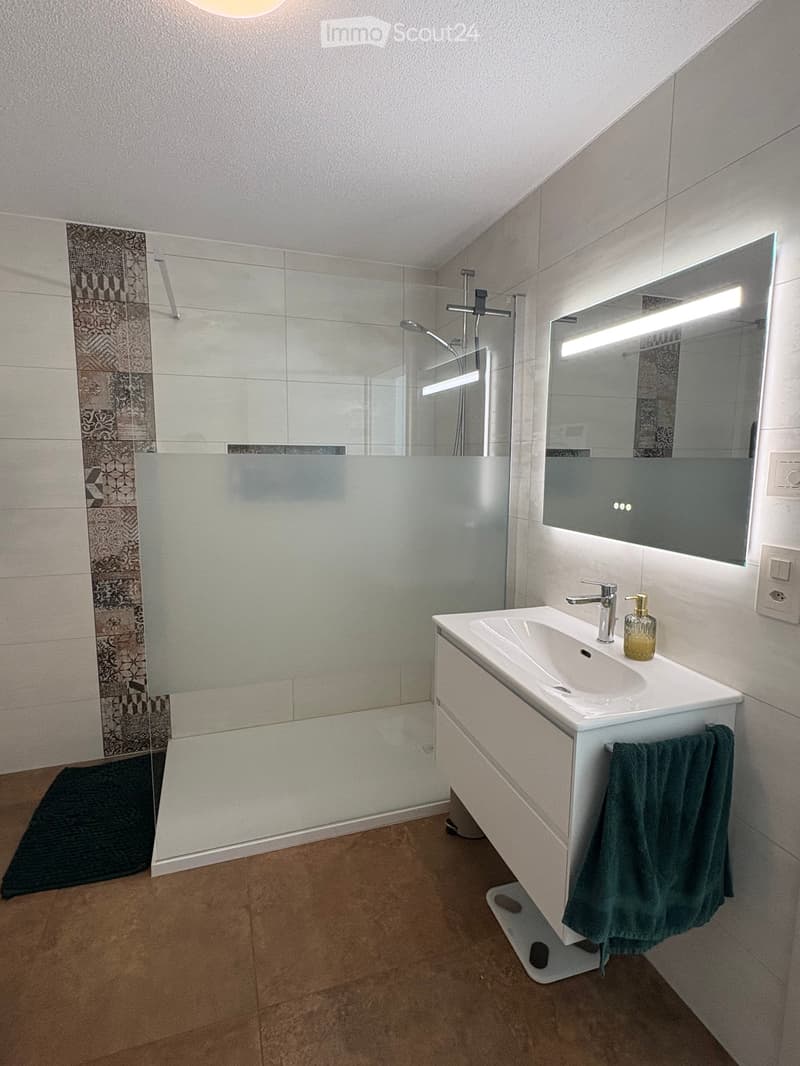 Modern bathroom with a white sink, chrome faucet, large mirror with built-in lighting, glass shower enclosure, and decorative tiles.