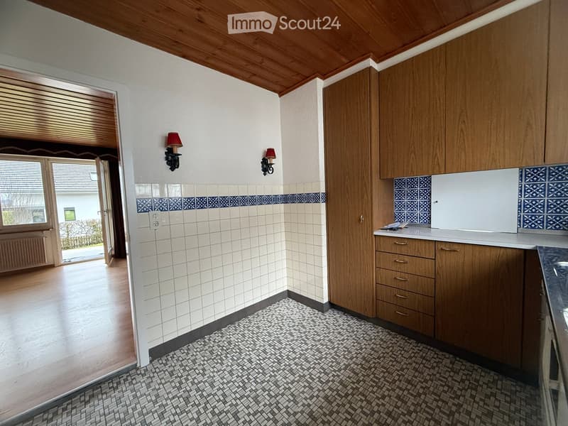 Wooden ceiling, white tiled wall, white and blue tiles on lower wall, wood cabinetry, white countertops, white and blue tiles on backsplash, wooden floor, open doorway, wall sconces, open window with a view of a house